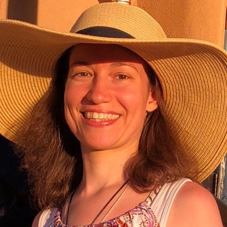 A headshot photo of Beth. She is weating a yellow, wide brimmed, woven hat with a black hat band at the base. Her brown hair hangs out from udnerneath the hat and touches her shoulders. Beth is facing the camera and smiling.